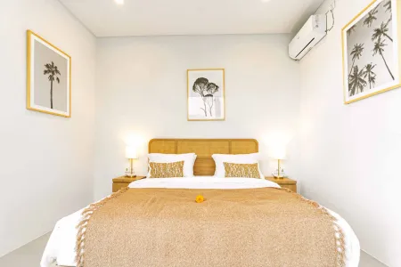 Cozy minimalist tropical bedroom with a wooden bed draped in beige linens, frangipani flower accent, palm tree wall art, and bedside lamps.